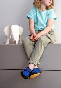 Niebieskie siatkowe buty z czarnymi akcentami i gumową podeszwą, noszone przez dziecko siedzące na szarym podłożu, obok białego obiektu w kształcie słonia.