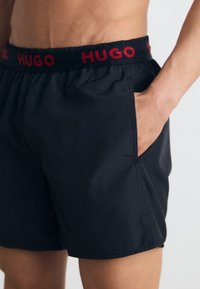 Čierne plávacie šortky s elastickým pásom, na ktorých je v červenej farbe nápis "HUGO", bočné vrecká, hladký povrch a dĺžka nad kolená.
