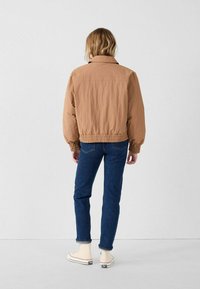 Giacca bomber marrone leggera con polsini ed orlo elasticizzati, abbinata a jeans blu e sneakers bianche, vista da dietro.