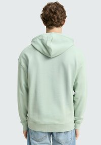 Uomo con capelli ricci che indossa una felpa con cappuccio verde chiaro e jeans blu, visto di spalle su uno sfondo semplice.