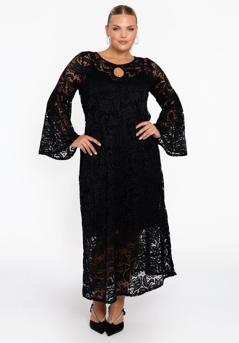 Robe en dentelle noire avec de longues manches en cloche et un décolleté en forme de clé. Motif floral transparent, conception longueur de sol, associée à des talons aiguilles noirs.