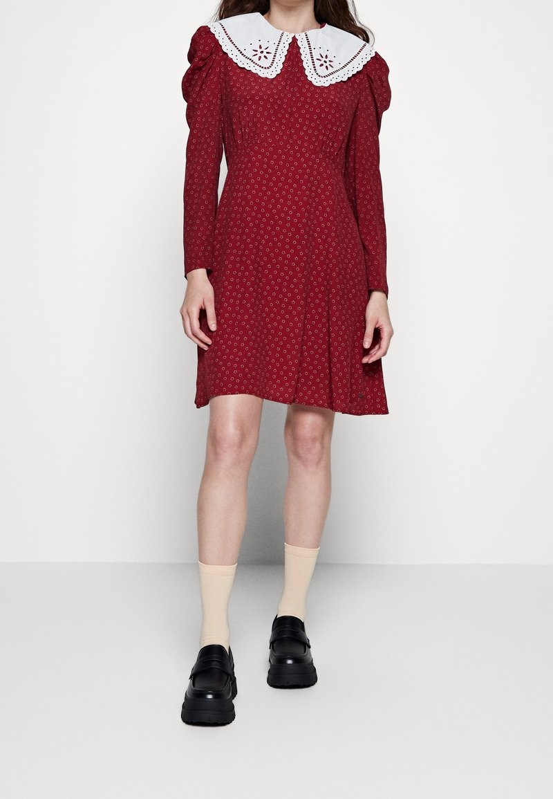 Femme portant une robe rouge à petits motifs floraux, col en dentelle blanche, chaussettes beige et chaussures noires épaisses, debout devant un fond blanc.