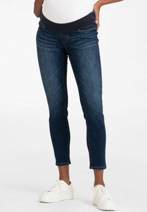 Person iført mørkeblå graviditetsjeans og hvide sneakers, stående med den ene hånd hvilende på hoften.