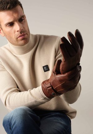 Gants en cuir marron avec une finition texturée et une sangle à boucle, portés par-dessus un pull ribbé beige clair, sur un fond neutre.