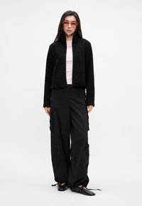 Svart fleecejakke med glidelås, rosa t-skjorte og svarte cargo-bukser. Svart sko med ankelremmer. Modell med klare solbriller.