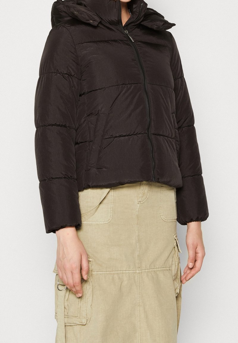 Doudoune noire avec fermeture éclair à l'avant et poignets élastiques portée sur une jupe cargo beige avec plusieurs poches et détail anneau.