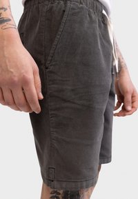 Κοντινό πλάνο ενός ατόμου που φορά σκούρα γκρι βερμούδα κοτλέ με μπροστινές τσέπες, λευκό κορδόνι και εμφανή τατουάζ στα χέρια και τα πόδια.