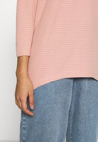 Helles rosa geripptes Oberteil mit Dreiviertelärmeln, kombiniert mit lockeren hellblauen Denim-Jeans. Detail ist eine leicht abgerundete Saumlinie.