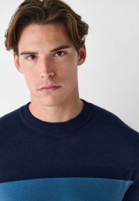 Tengerészkék kötött pulóver vízszintes világoskék csíkkal, kerek nyakkivágással és puha tapintású anyagból. Közeli kép a felsőtest és az arc részleteiről.