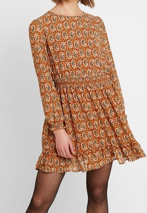 Frau trägt ein langärmeliges, knielanges oranges Kleid mit Paisley-Muster und Rüschen am Saum, kombiniert mit transparenten schwarzen Strumpfhosen.