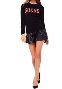 Schwarzer Pullover mit pinkem "GUESS"-Logo, kombiniert mit glänzenden schwarzen Shorts und schwarzen High Heels. Fokus auf lässigen Stil und kontrastierende Texturen.