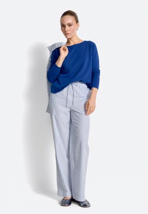 Woman wearing a blue long-sleeve top tucked into high-waisted, light blue striped wide-leg pants, holding a matching striped jacket.