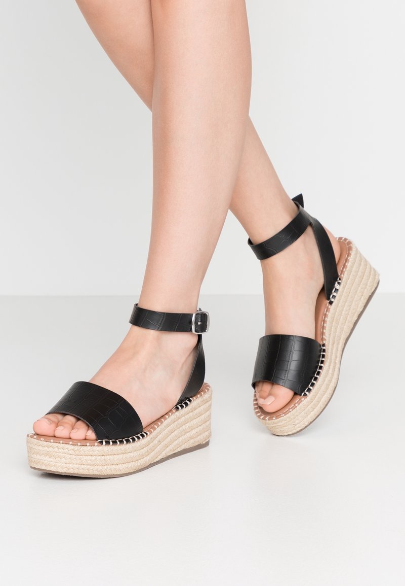 Sandales compensées en cuir noir avec une texture en peau de crocodile. Elles présentent une large sangle, une boucle à la cheville et une plateforme tressée en jute.
