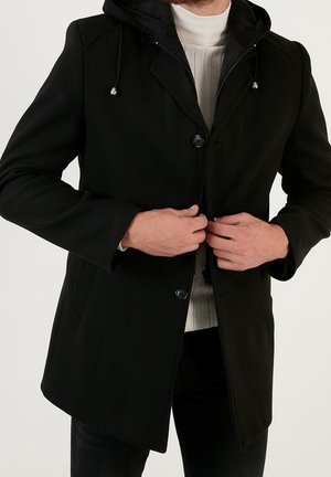 Man wearing black hooded coat over white sweater, fastening the coat's buttons and zipper.