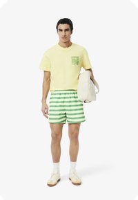 Camiseta amarilla con bolsillo gráfico verde, pantalones cortos a rayas verdes y blancas, y bolsa de tela beige. Zapatillas blancas realzan el look casual.