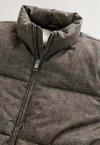 Käytössä oleva harmaa tilkkuvuorattu takki korkealla kauluksella, vetoketjulla ja tekstuurilla. Sisältää hienovaraisen ruudullisen kuvion ja siistit saumat.