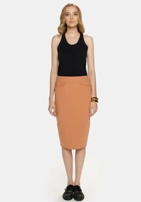Czarna bezrękawnikowa bluzka połączona z dopasowaną, sięgającą kolan, jasnobrązową spódnicą z dwiema przednimi kieszeniami, noszona w komplecie z czarnymi butami.