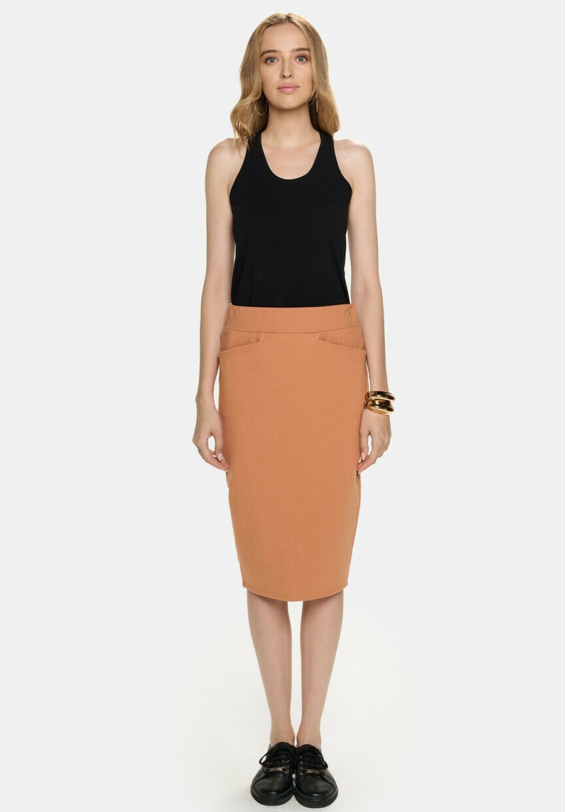 Czarna bezrękawnikowa bluzka połączona z dopasowaną, sięgającą kolan, jasnobrązową spódnicą z dwiema przednimi kieszeniami, noszona w komplecie z czarnymi butami.
