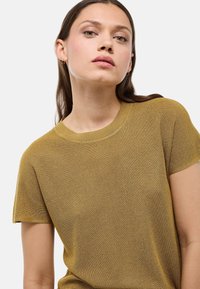 Femme à la peau claire et aux cheveux bruns portant des boucles d'oreilles en or en forme de créole et un haut en tricot jaune moutarde à manches courtes, sur fond uni clair.