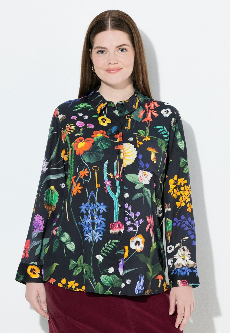 Donna che indossa una camicia nera con bottoni decorata con motivi floreali colorati, cactus, funghi e chiavi, abbinata a una gonna rosso scuro.