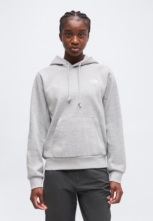 Jeune personne aux cheveux tressés portant un sweat à capuche gris clair et un pantalon noir, debout devant un fond blanc uni.