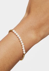 Pulsera de perlas con perlas blancas y cuentas con detalles dorados, ensartadas en una banda elástica, llevada en una muñeca.