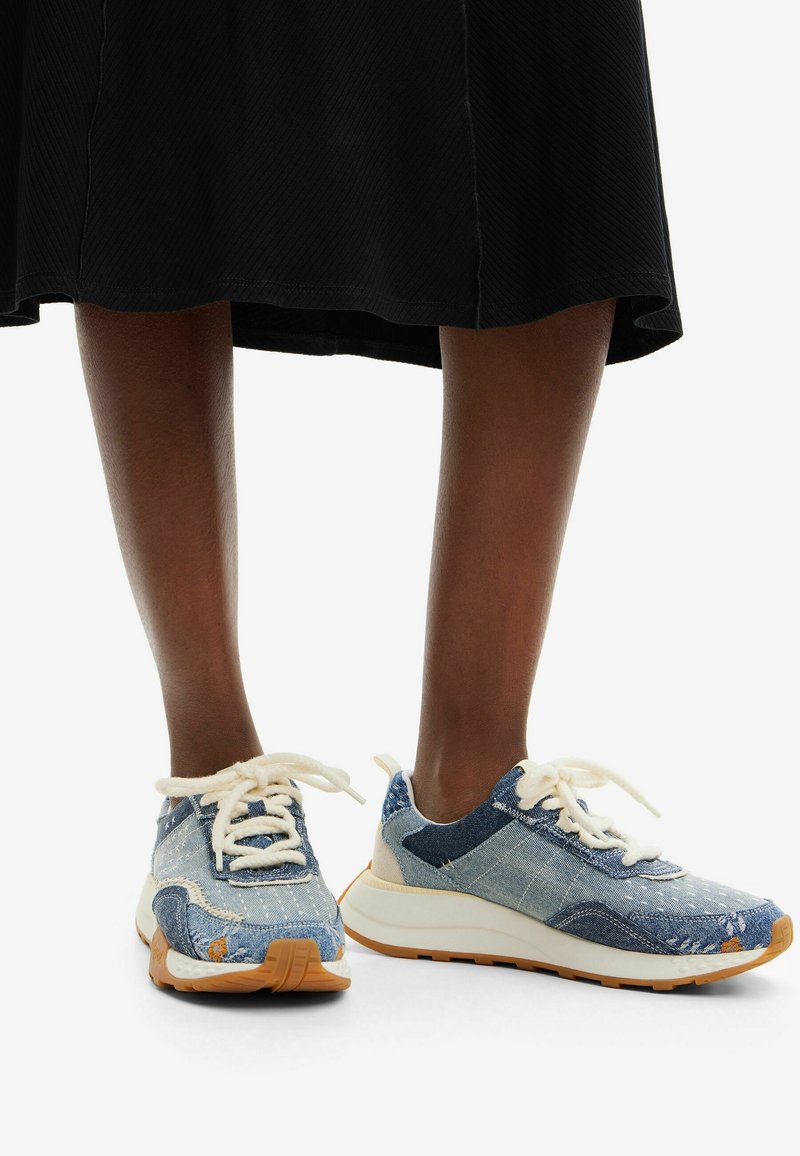Jambes portant une jupe noire et des baskets bleues patchwork avec des lacets blancs et des semelles épaisses blanches et marron, debout sur une surface blanche.