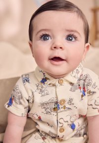 Īsām piedurknēm krēmkrāsas kombinezons ar košiem cirka tematikas apdrukām, pogām un salocītu apkakli, izgatavots no mīksta, teksturēta auduma.