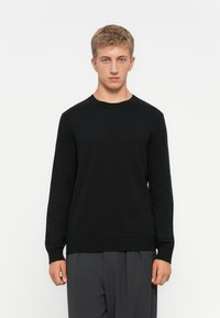 Young man in black long-sleeve sweater and gray pants standing against white background, facing forward with neutral expression.