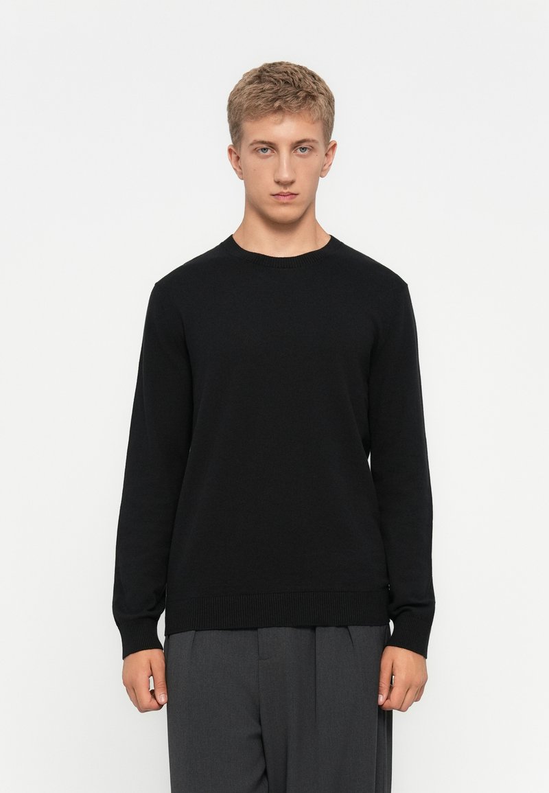Young man in black long-sleeve sweater and gray pants standing against white background, facing forward with neutral expression.