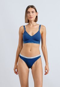 Žena stoji, nosi usklađene tamnoplave bralette i donje rublje Calvin Klein s elastičnim trakama s logotipom, neutralnog izraza, svijetla pozadina.