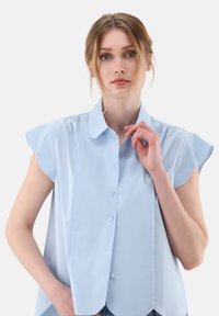 Blusa azzurra a maniche corte con colletto a punta, chiusura frontale con bottoni e dettagli arricciati sulle spalle. Tessuto morbido.