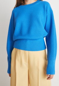 Femme portant un pull bleu texturé à manches longues rentré dans un pantalon beige taille haute à jambes larges, sur un fond uni.