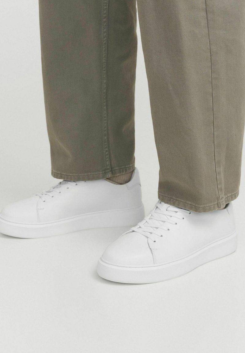 Baskets en cuir blanc avec une texture lisse, bout arrondi et semelle en caoutchouc. Portées avec un pantalon beige clair, doté d'une coupe droite.