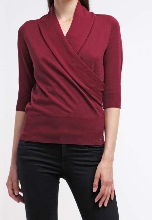 Femme portant un pull ajusté style cache-cœur bordeaux avec manches trois-quarts et pantalon noir sur un fond blanc uni.