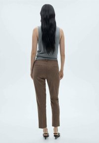 Haut gris sans manches, pantalon à carreaux marron avec une coupe slim, vue arrière. Sandales à talons noirs et cheveux lisses. Arrière-plan clair.