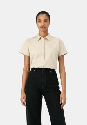 Donna che indossa una camicia beige a maniche corte con bottoni infilata in pantaloni neri a vita alta, in piedi davanti a uno sfondo chiaro e uniforme.
