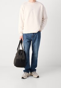 Persona che indossa un maglione bianco sporco, jeans blu, sneakers beige, tiene una borsa a tracolla nera e grigia con la mano sinistra, mentre la mano destra è in tasca.