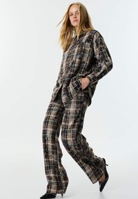 Completo composto da camicia e pantaloni a fantasia scozzese nei toni del marrone, beige e verde. Il materiale ha una consistenza morbida, con tasche e una vestibilità rilassata. Le scarpe nere aggiungono contrasto.