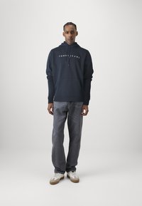Felpa navy con logo "TOMMY JEANS", tasca frontale, polsini a coste, abbinata a jeans dritti grigio chiaro e sneakers bianche con dettagli color marrone.