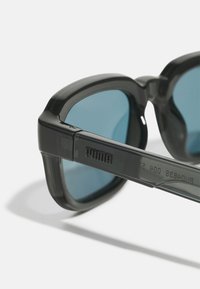 Ochelari de soare cu un cadru gros, din plastic semi-transparent de culoare gri închis și lentile de culoare albastră. Prezintă marca PUMA pe braț.