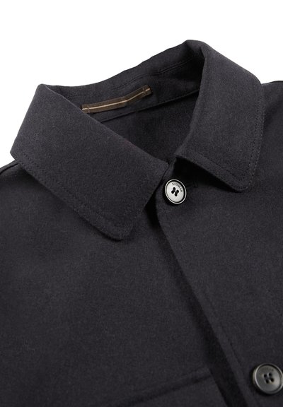 Cappotto di lana nera con colletto a punta, chiusura con bottoni e una texture sottile. Hardware notevole include un bottone con logo argentato.