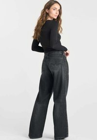 Vrouw met lang bruin haar, gekleed in een zwarte lange mouwtop en hoge taille wijde grijze jeans, kijkt over haar schouder.