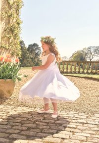 Ragazzina con corona di fiori che gira in un vestito bianco e scarpe su un sentiero di ciottoli assolato vicino a un vaso di tulipani.