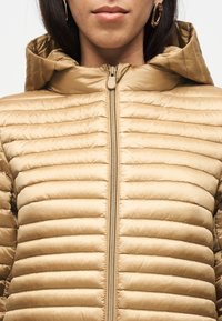 Γυναίκα που φοράει μπεζ καπιτονέ μπουφάν με κουκούλα και φερμουάρ κλειστό, με εμφανή χρυσά σκουλαρίκια κρίκους, πάνω σε απλό φόντο.