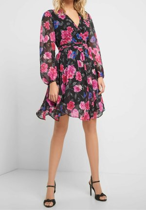 Vrouw in een zwart bloemenjurk tot op de knie met lange doorschijnende mouwen en zwarte sandalen met hoge hakken, staand tegen een effen witte achtergrond.