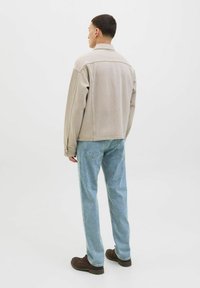 Jachetă de denim bej, croi relaxat, cusături vizibile, guler înalt; asociată cu blugi de culoare bleu deschis și pantofi maro închis. Fundal simplu.
