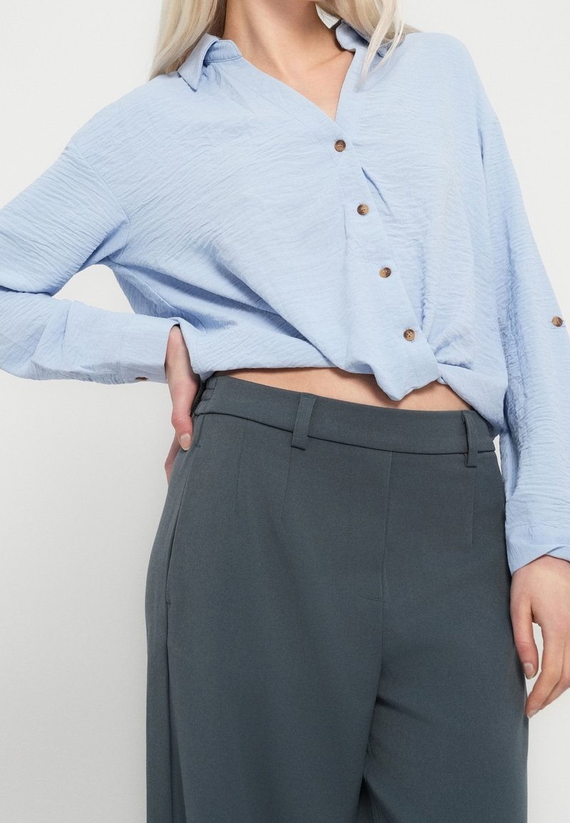 Femme portant un chemisier bleu clair boutonné, légèrement rentré dans un pantalon gris foncé taille haute, une main posée sur la hanche.