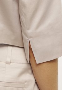 Chemise beige à manches courtes avec fente latérale portée sur un pantalon beige avec passants de ceinture et détails de poche arrière.