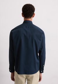 Man met kort krullend haar, gekleed in een marineblauw overhemd met lange mouwen en beige broek, gezien van achteren tegen een effen achtergrond.